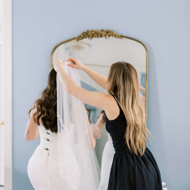 Stylist putting a veil on a bride who is wearing an aisle-ready bridal gown in front of a mirror at Bleu bridal Co